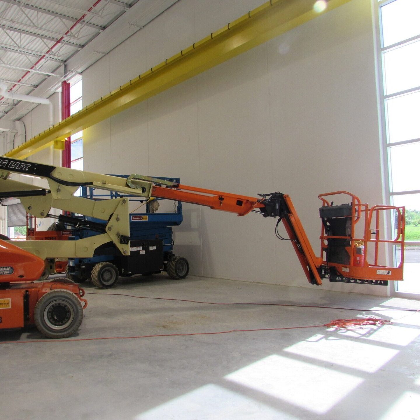 Orange boom lift extended near windows in a warehouse or industrial building. Blue scissor lift in background.